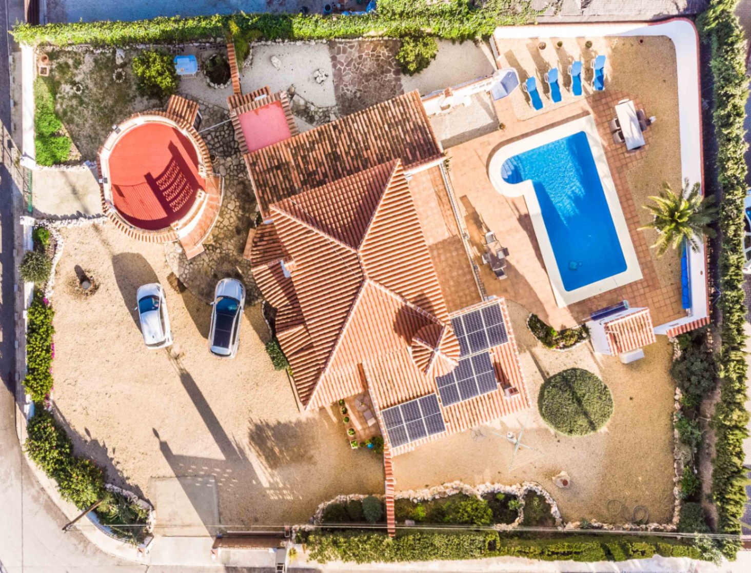Vista aérea cenital que muestra la distribución de la parcela, la piscina en forma de L, los paneles solares sobre el tejado y la singular torre circular.