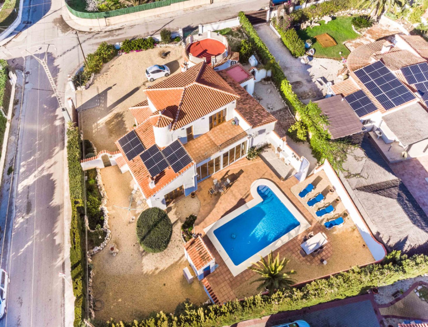 Vista aérea de la villa con tejado de teja, paneles solares y piscina privada rodeada de zonas de descanso en una parcela vallada con setos.