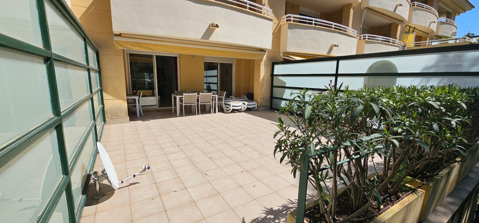 Amplia terraza privada con suelo cerámico, toldos extensibles amarillos y zona de comedor exterior protegida por paneles de vidrio translúcido y vegetación.