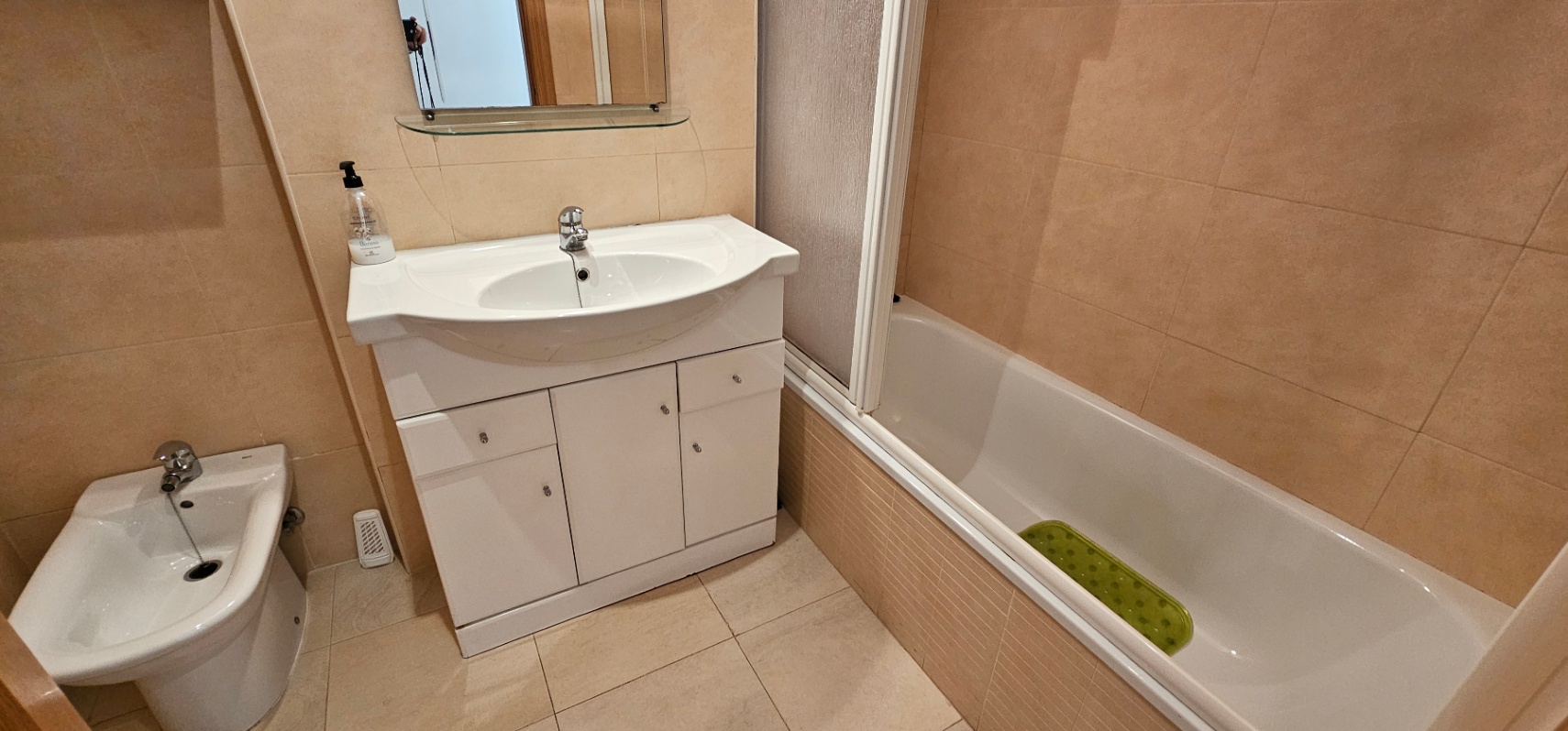 Baño completo con revestimiento cerámico en tonos beige, mueble de lavabo blanco con almacenaje, bidé y bañera con mampara.