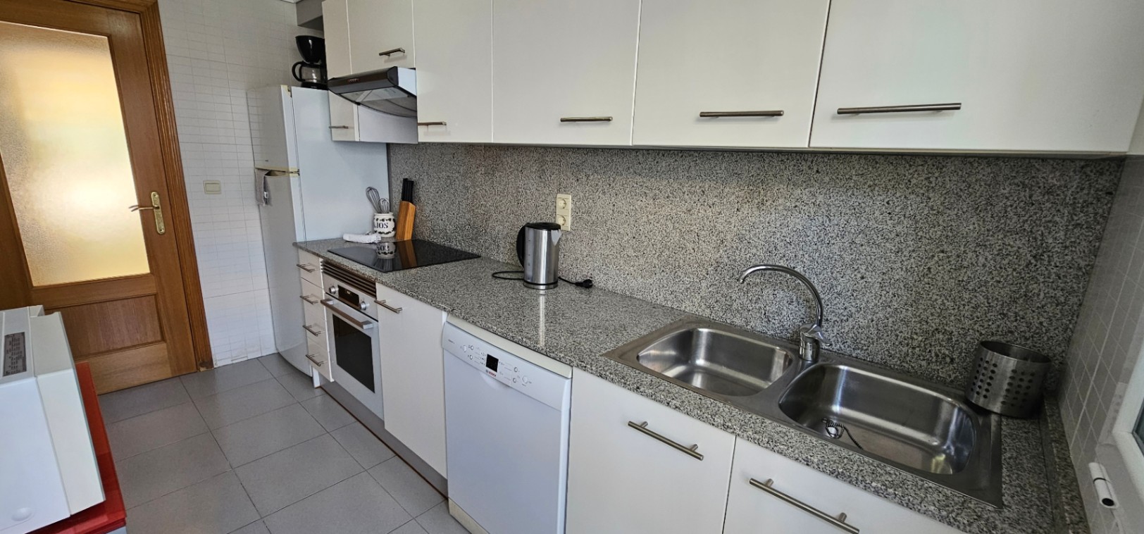 Cocina funcional con encimera y frontal de granito gris, armarios blancos, electrodomésticos integrados y fregadero de doble seno en acero inoxidable.