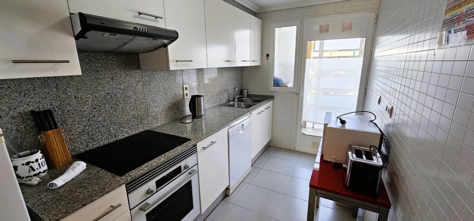 Cocina independiente equipada con encimera de granito, muebles blancos y electrodomésticos integrados. Incluye acceso directo a una galería exterior con luz natural.