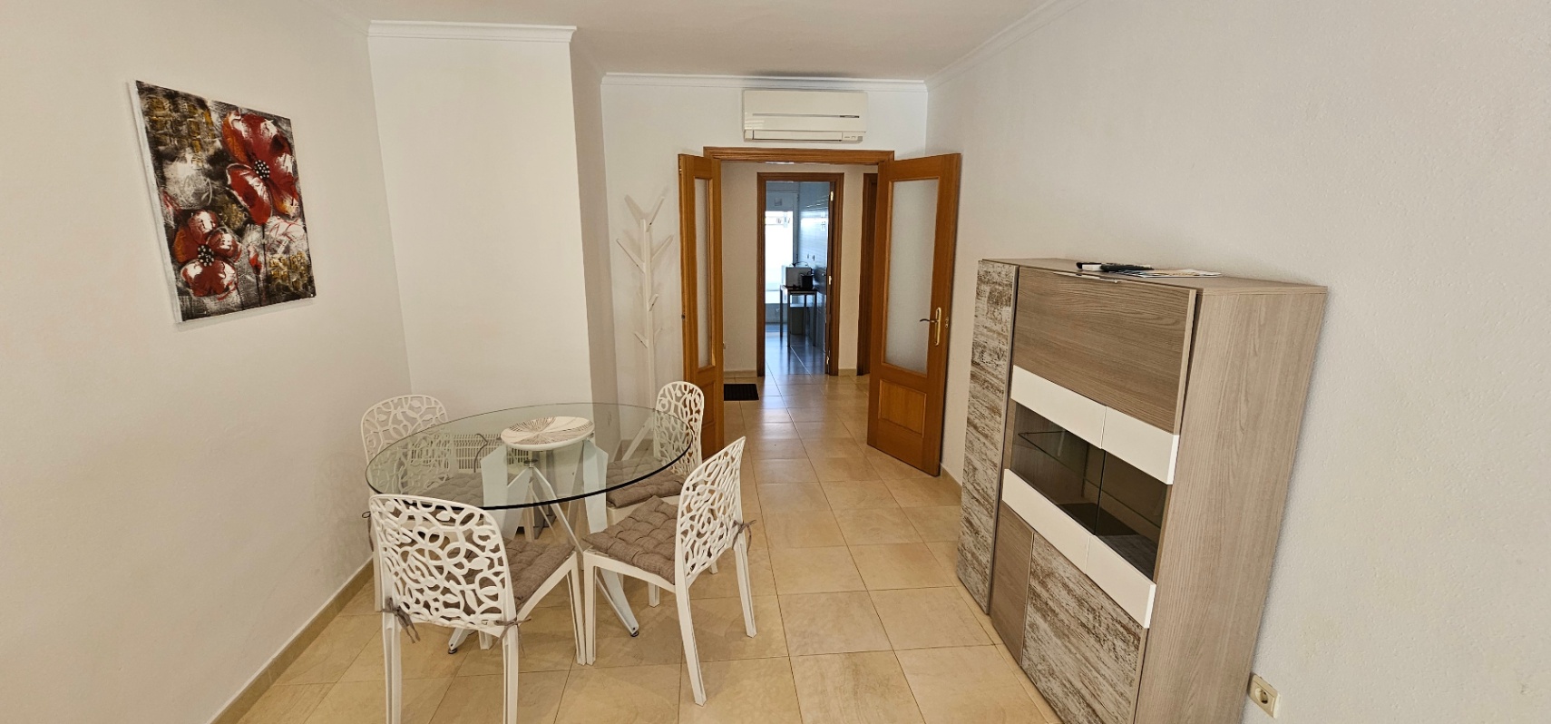 Comedor con mesa circular de cristal, sillas blancas de diseño y aparador moderno. Incluye suelos cerámicos, aire acondicionado y puertas dobles de madera.