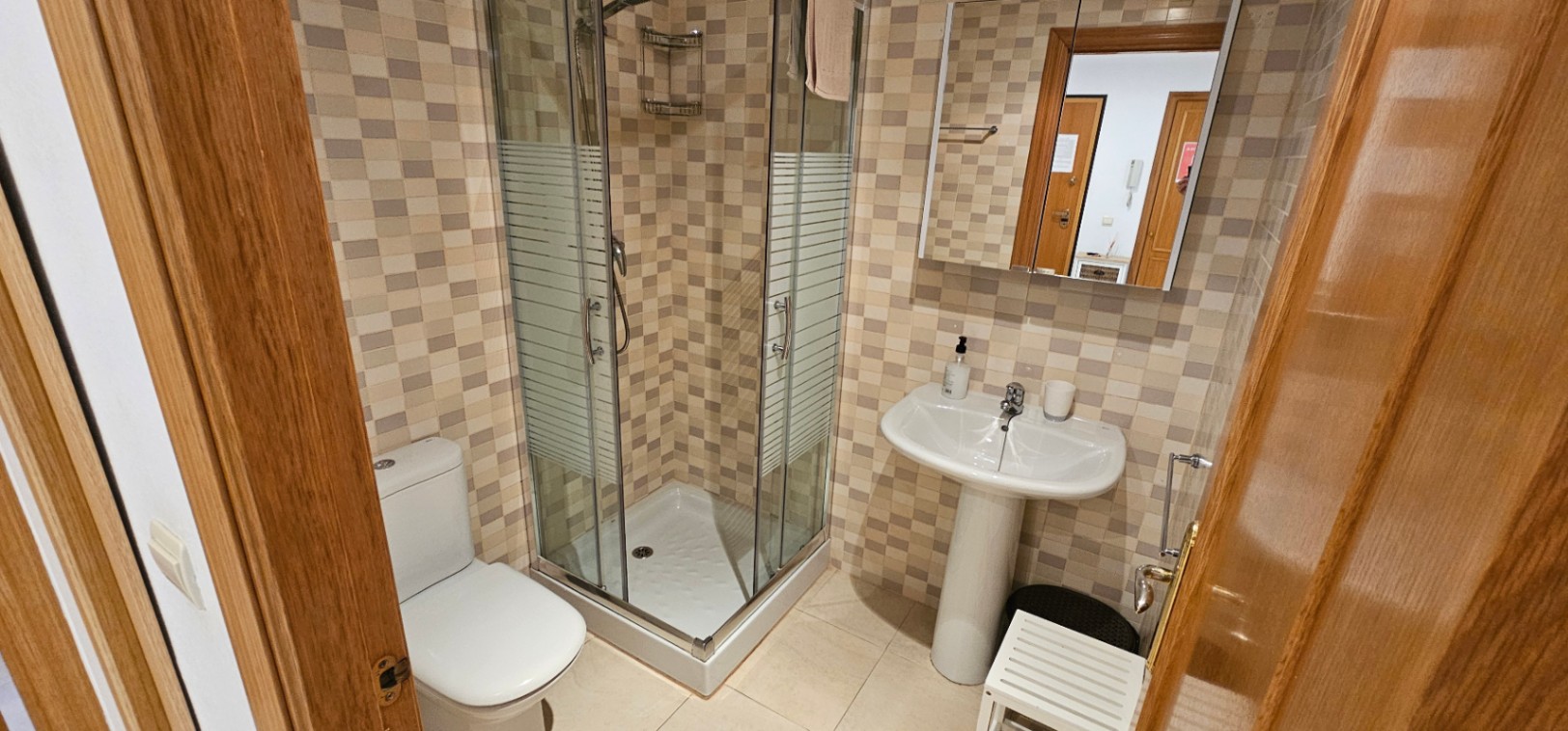 Cuarto de baño con ducha de rincón con mampara de cristal, lavabo de pedestal y paredes revestidas con azulejos de mosaico en tonos tierra.