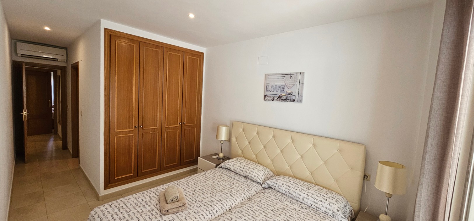 Dormitorio con armario empotrado de madera de cuatro hojas, cabecero acolchado en tono crema y aire acondicionado integrado sobre el acceso al pasillo.