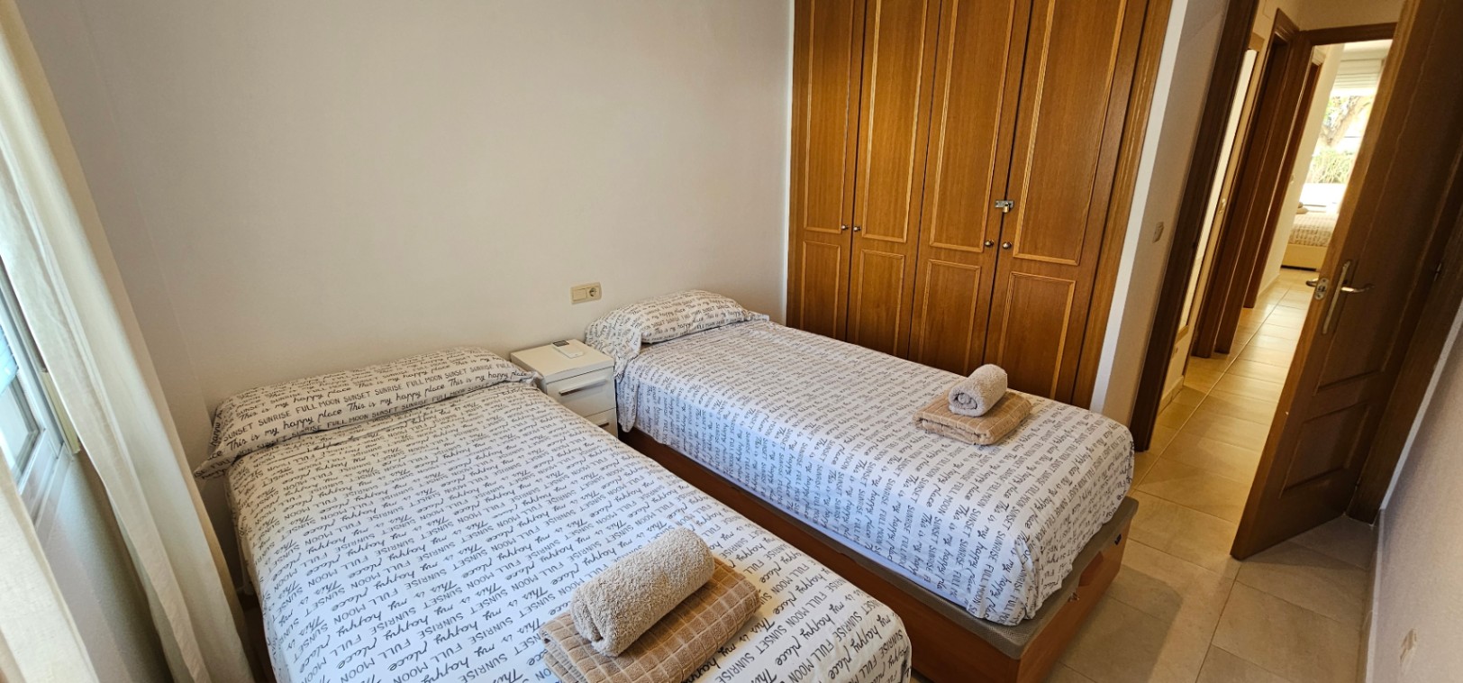Dormitorio con dos camas individuales, amplio armario empotrado de madera y suelos de gres, iluminado por luz natural lateral.