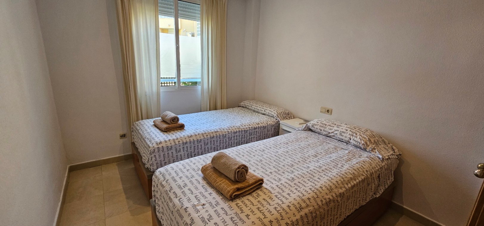 Dormitorio con dos camas individuales, suelos de gres claro y ventana exterior con persiana y cortinas blancas.