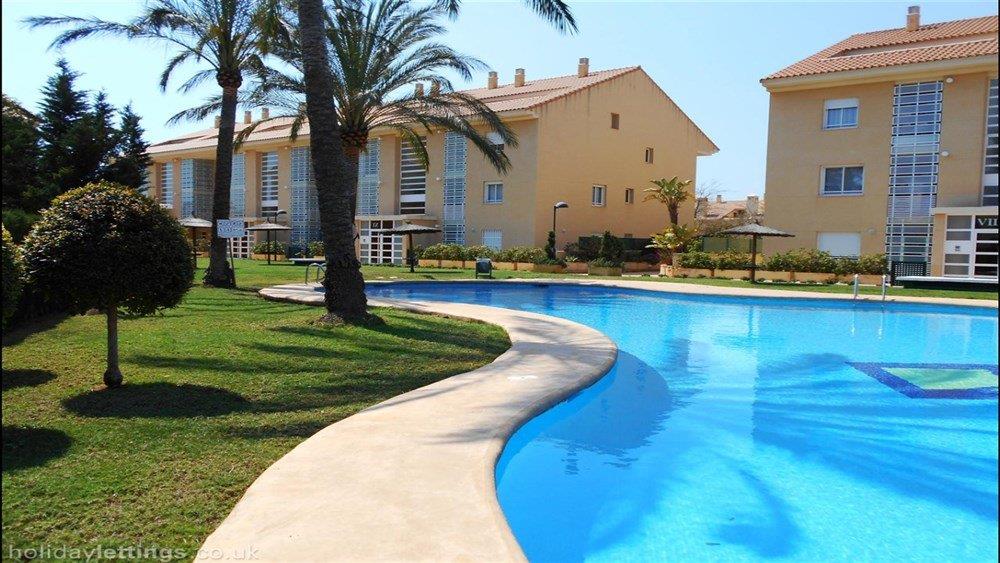 Piscina comunitaria de diseño curvo rodeada de amplias zonas verdes, palmeras y edificios residenciales de estilo mediterráneo en Jávea.
