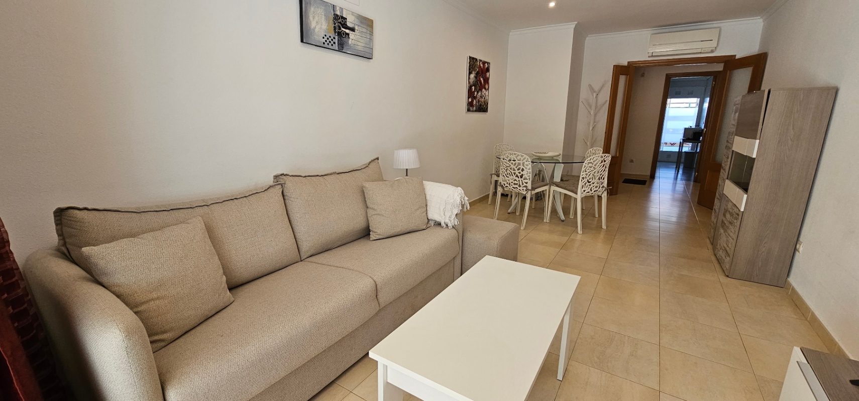 Salón-comedor luminoso con sofá beige, mesa de centro blanca y zona de comedor con sillas de diseño. Incluye aire acondicionado y suelos cerámicos de tonos neutros.