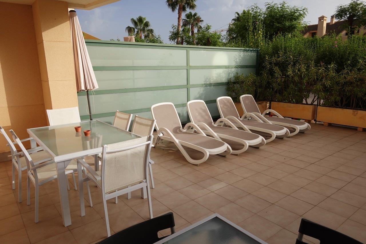 Terraza privada con suelo cerámico, equipada con zona de comedor exterior y tumbonas, delimitada por pantallas de cristal translúcido y vegetación perimetral.