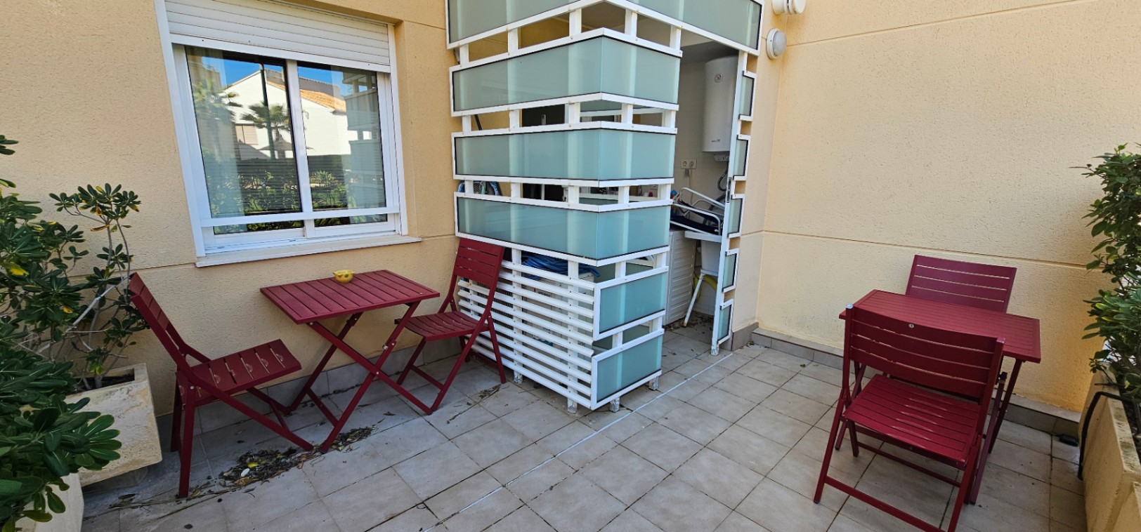 Terraza privada con suelo cerámico, mobiliario exterior de madera rojiza y un práctico cerramiento de cristal translúcido para la zona de lavadero.