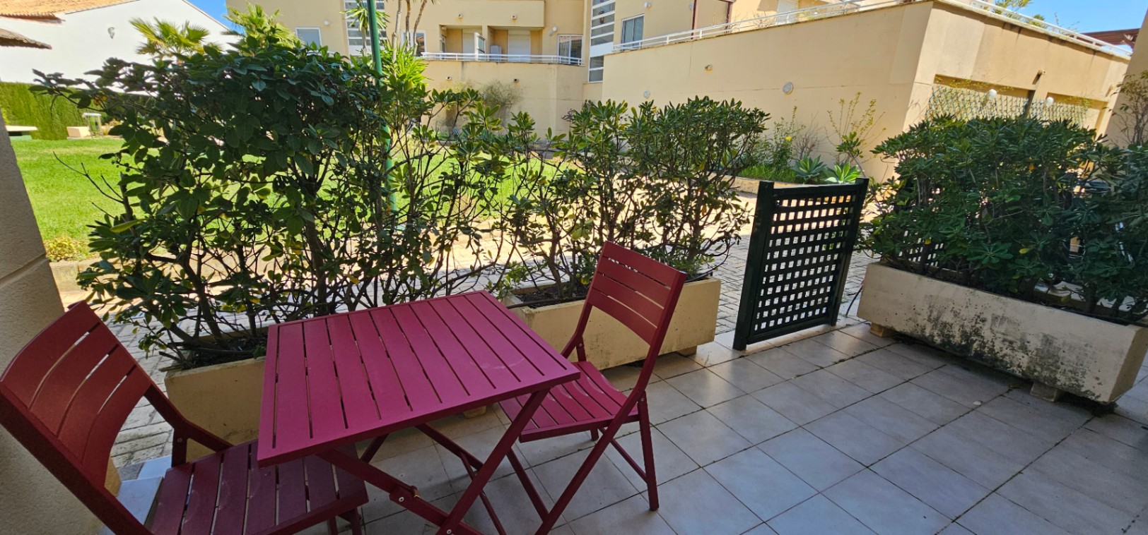 Terraza privada con suelo de baldosas claras, mobiliario de exterior en color granate y acceso directo a las zonas ajardinadas de la urbanización.
