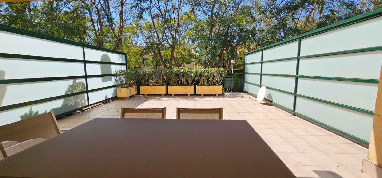 Terraza privada con suelo de gres, paneles laterales de vidrio traslúcido para mayor privacidad y jardineras con arbustos bajo árboles maduros.