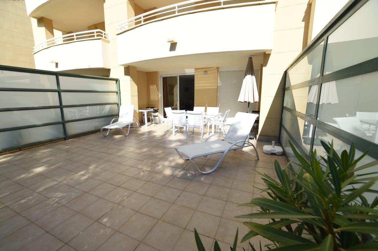 Terraza privada de planta baja con suelo cerámico, equipada con tumbonas blancas y zona de comedor exterior bajo el porche del edificio.
