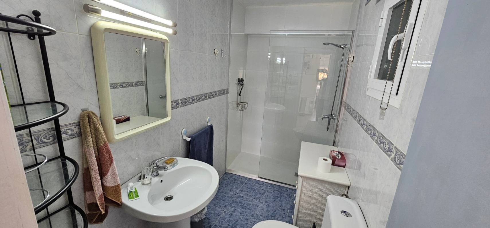 Baño funcional con ducha de obra acristalada, lavabo de pedestal y suelos cerámicos azules. Incluye azulejos claros con cenefa decorativa y ventana para ventilación natural.