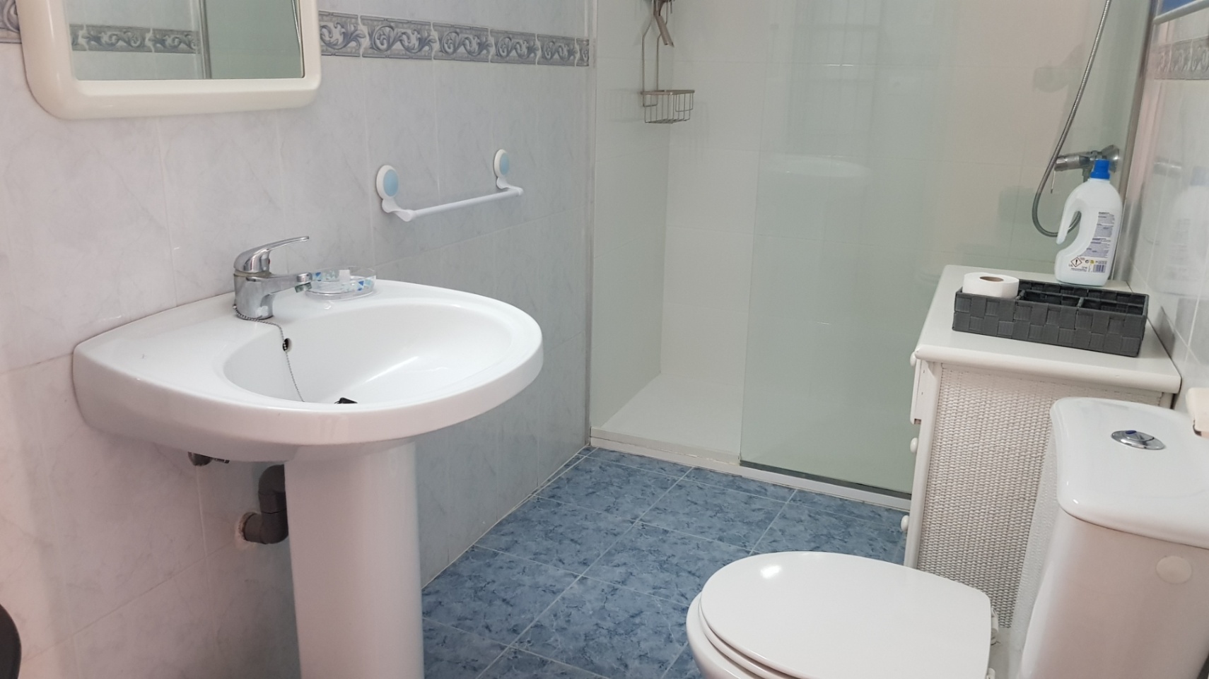 Baño funcional con lavabo de pedestal, inodoro y ducha con mampara de cristal. Presenta un suelo cerámico azul y un mueble de almacenamiento en mimbre blanco.