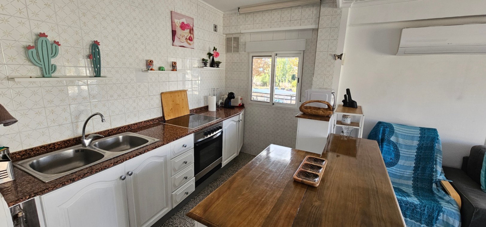 Cocina de planta abierta con encimera de granito, fregadero doble y armarios blancos. Incluye mesa de comedor de madera, aire acondicionado y ventana al exterior.