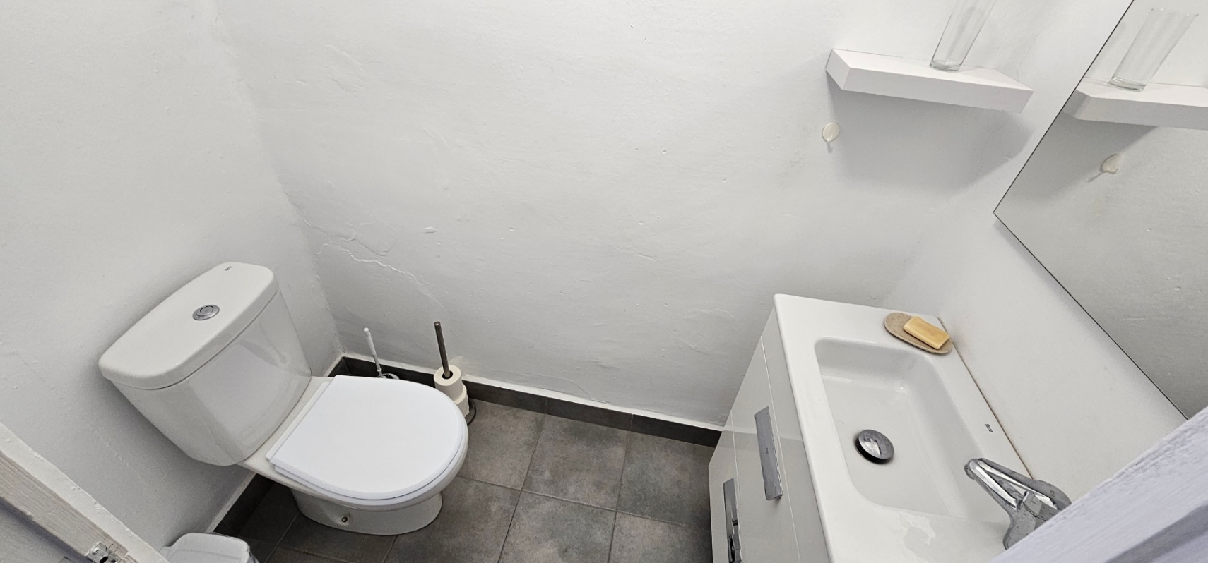 Cuarto de baño funcional con inodoro blanco, lavabo integrado en mueble de alto brillo y suelo de baldosas grises. Incluye un espejo y un estante de pared minimalista.
