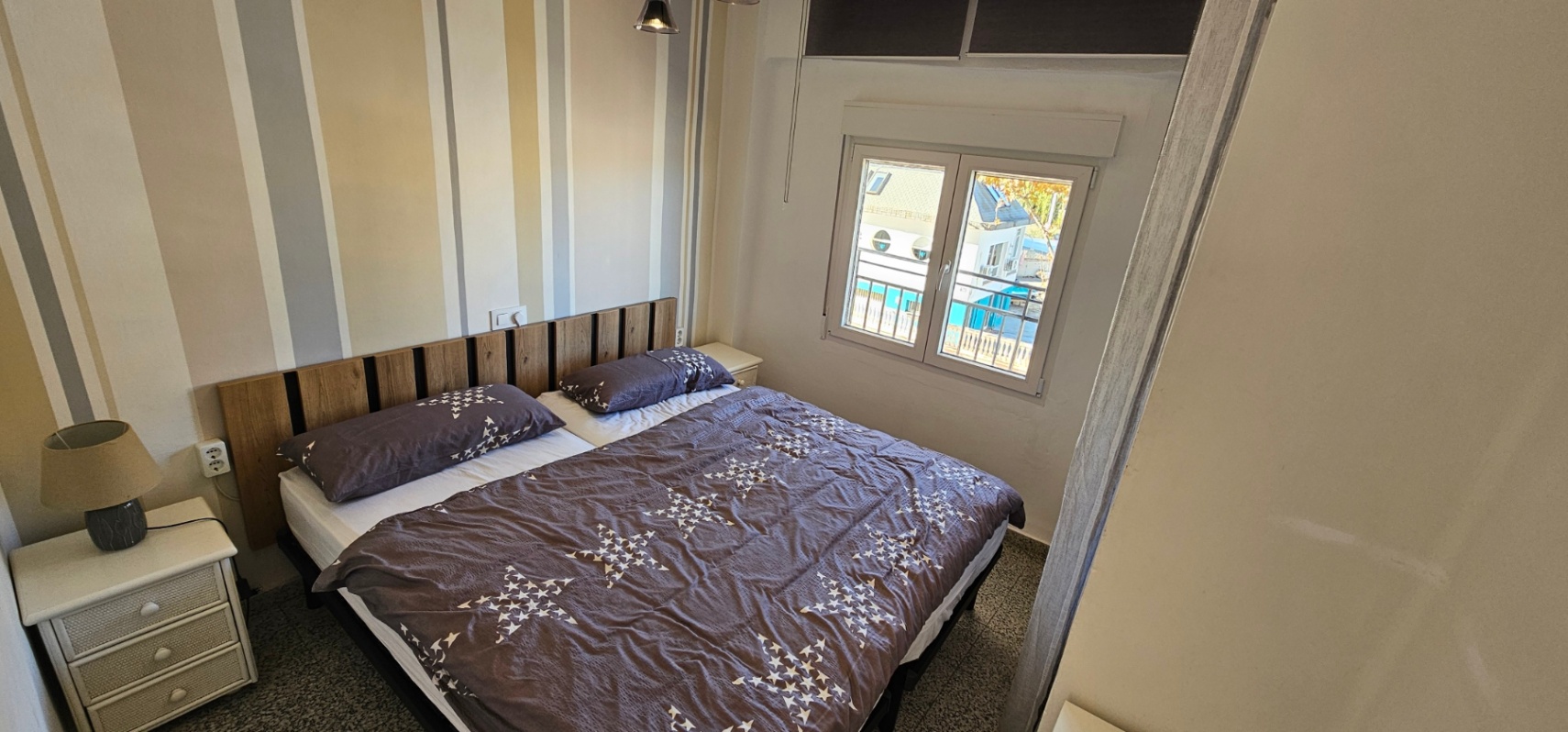 Dormitorio con cama doble, cabecero de listones de madera y papel pintado a rayas. Incluye ventana exterior que aporta luz natural y mesitas de noche blancas.