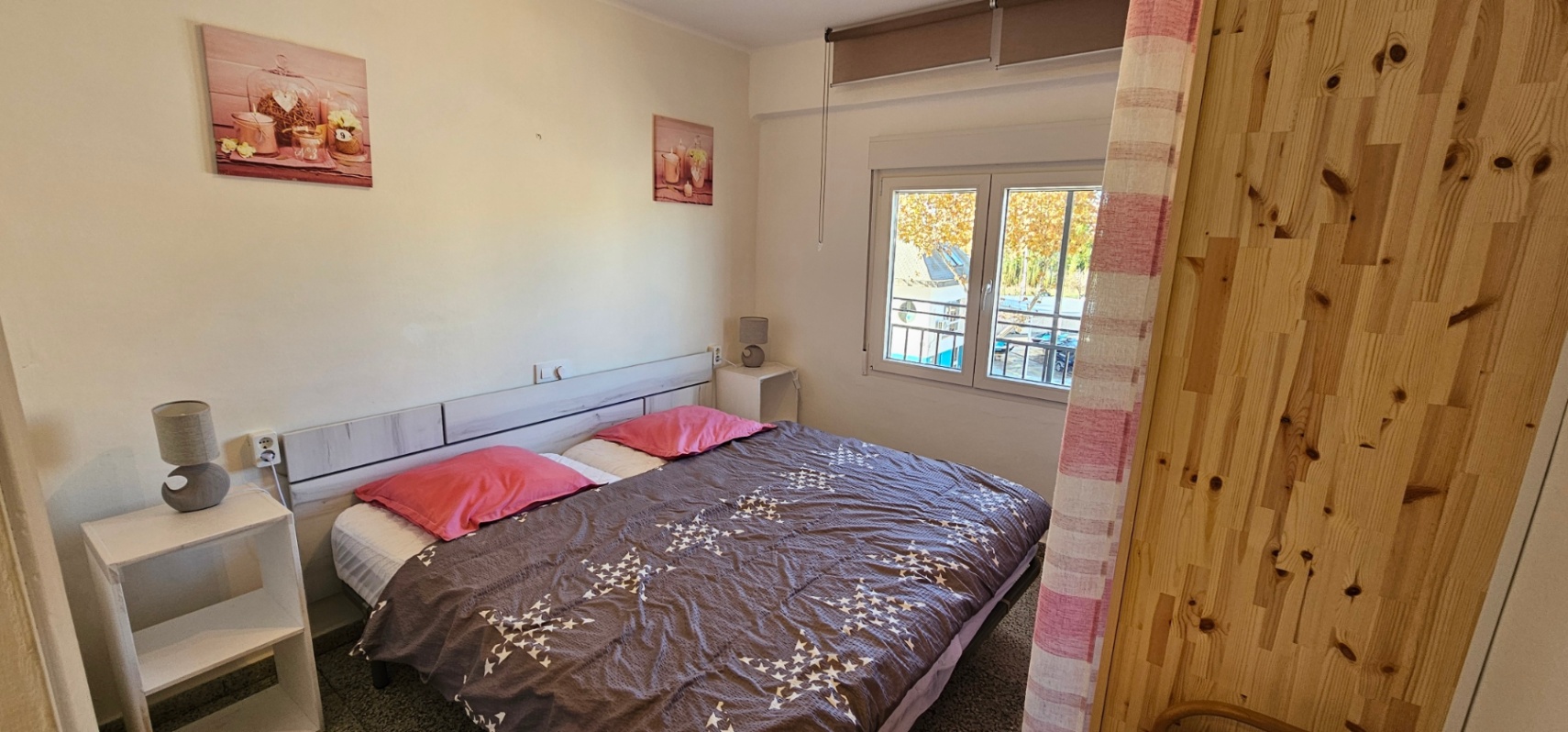 Dormitorio con cama doble, cabecero de madera clara y mesitas blancas. Incluye ventana exterior con persiana y vistas al entorno, aportando luz natural a la estancia.