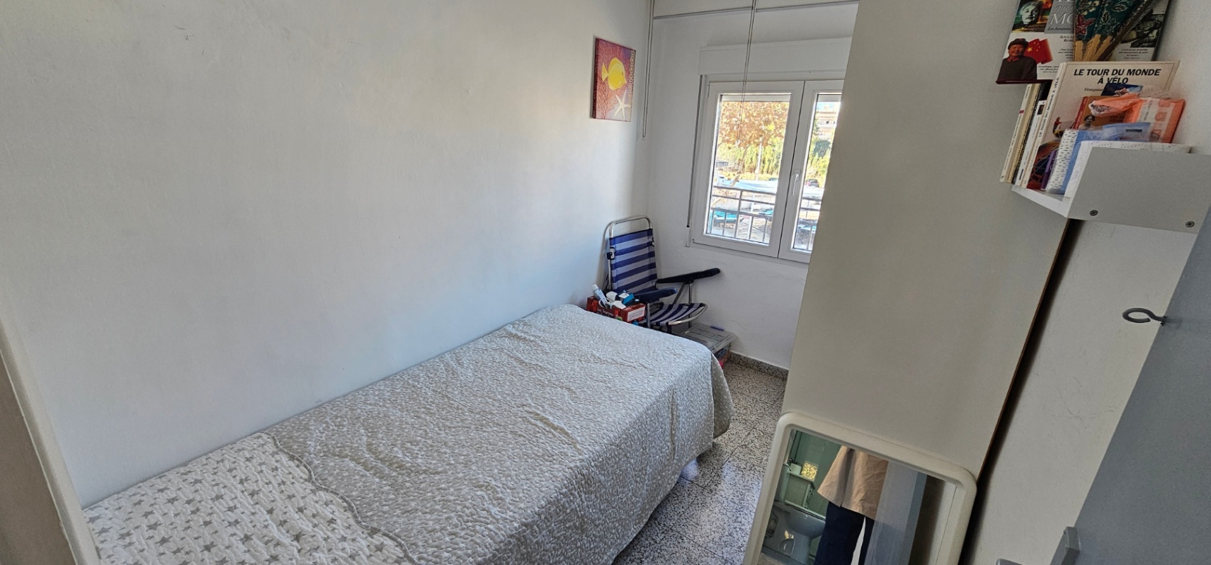 Dormitorio individual con suelos de terrazo, cama con colcha estampada y ventana exterior que permite la entrada de luz natural.