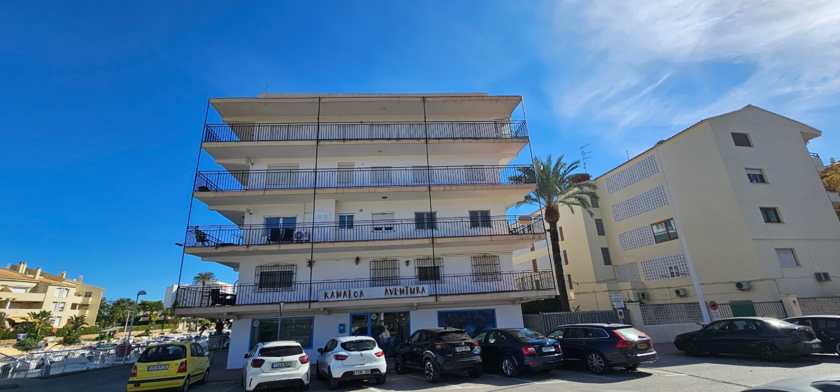 Fachada exterior del edificio de estilo mediterráneo con amplias terrazas perimetrales y barandillas metálicas, situado frente a una zona de aparcamiento.