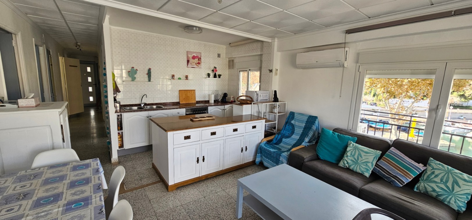 Salón-comedor de planta abierta con cocina integrada, isla central de madera y acceso directo a la terraza con vistas al canal y las embarcaciones.