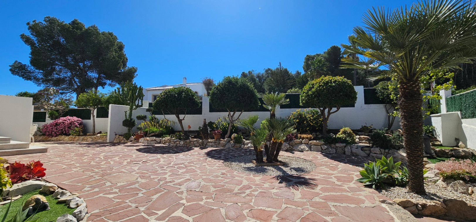 Amplio patio exterior con pavimento de piedra irregular en tonos rojizos, rodeado de parterres con vegetación mediterránea, palmeras y muros blancos bajo un cielo despejado.