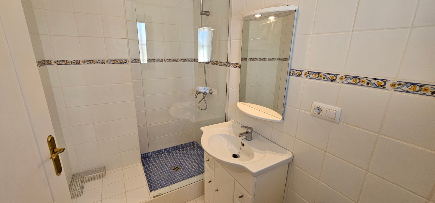Baño funcional con ducha de obra, suelo de mosaico azul, mampara de cristal y lavabo con mueble blanco y espejo iluminado.