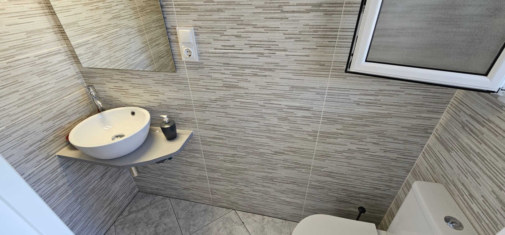 Baño funcional con lavabo de sobremesa circular, encimera suspendida gris y paredes revestidas con azulejos de textura lineal en tonos neutros.