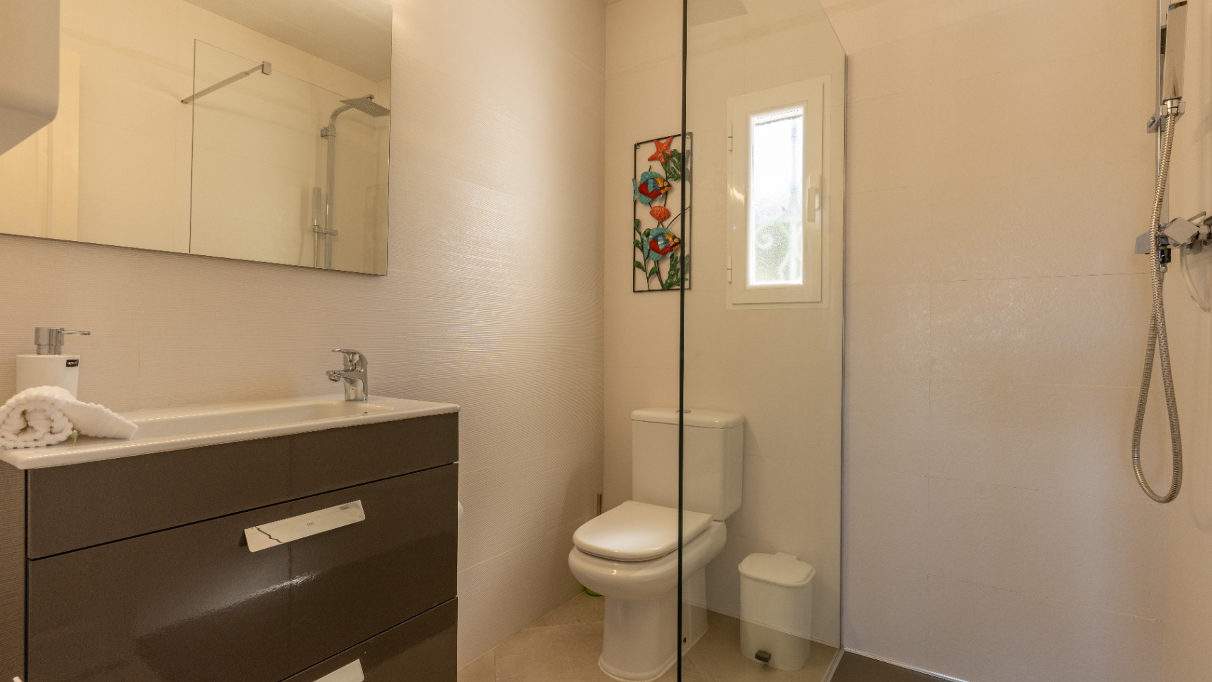 Baño moderno con mueble de lavabo en tono gris oscuro, mampara de ducha de cristal y azulejos en tonos crema con textura lineal sutil.