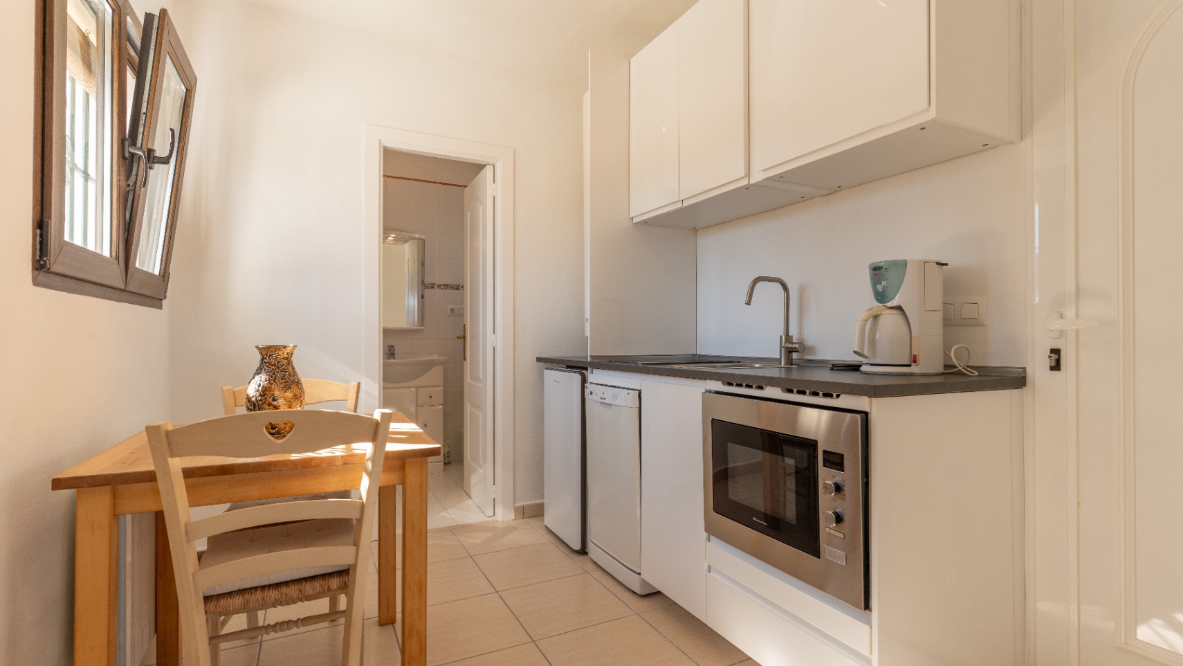 Cocina funcional con armarios blancos, encimera oscura y electrodomésticos integrados. Incluye una zona de comedor con mesa de madera y acceso directo a un baño.