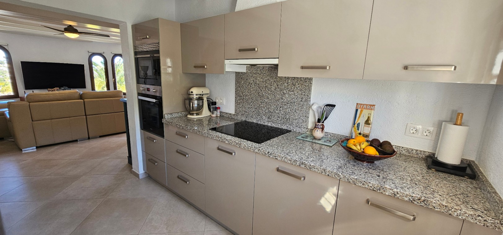 Cocina independiente con armarios de acabado brillo en tono topo, encimera de granito y electrodomésticos integrados, conectada visualmente con el salón.