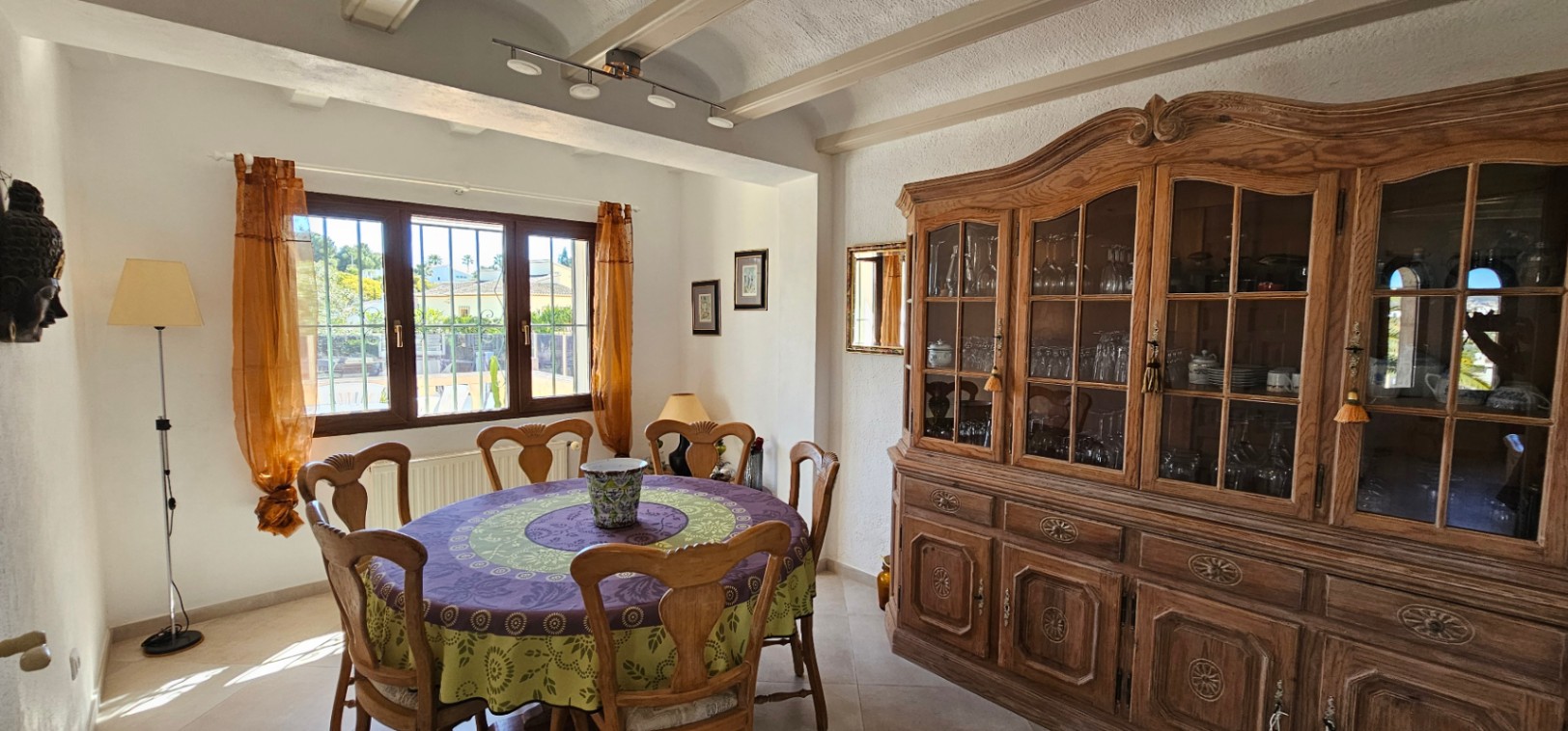 Comedor con mesa redonda, sillas de madera tallada y una gran vitrina clásica. Destacan los techos con vigas vistas y la entrada de luz natural por el ventanal.