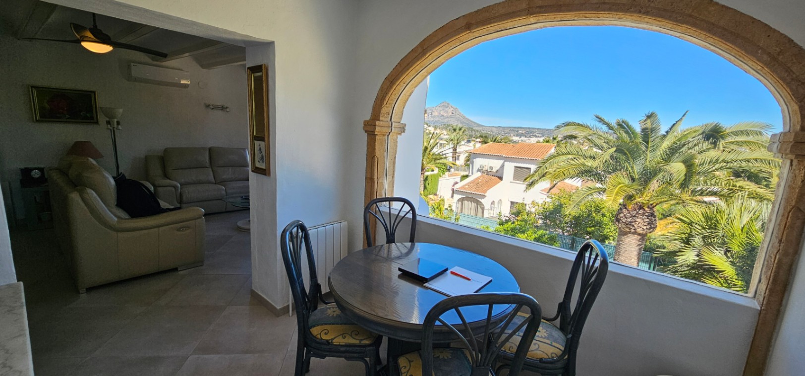 Comedor en naya con arco de piedra tosca y vistas al Montgó. El espacio conecta con un salón interior equipado con sofás de piel y ventilador de techo.