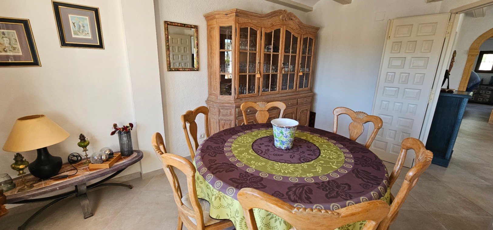 Comedor rústico con mesa circular, sillas de madera tallada y una amplia vitrina clásica. La estancia cuenta con suelos cerámicos y luz natural indirecta.