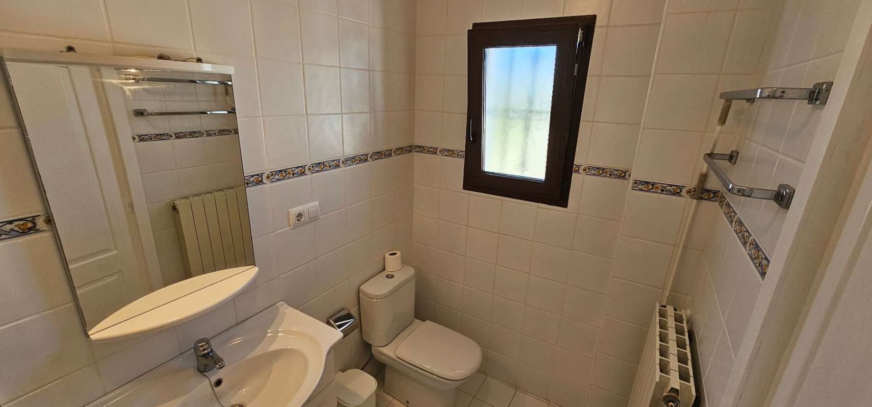 Cuarto de baño con azulejos blancos y cenefa decorativa, equipado con inodoro, lavabo con espejo y ventana de marco oscuro para ventilación natural.