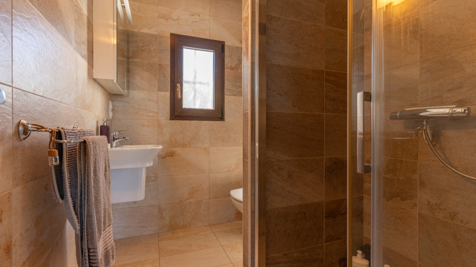 Cuarto de baño con revestimiento cerámico de efecto piedra, ducha con mampara de cristal, lavabo blanco y ventana con marco de madera.