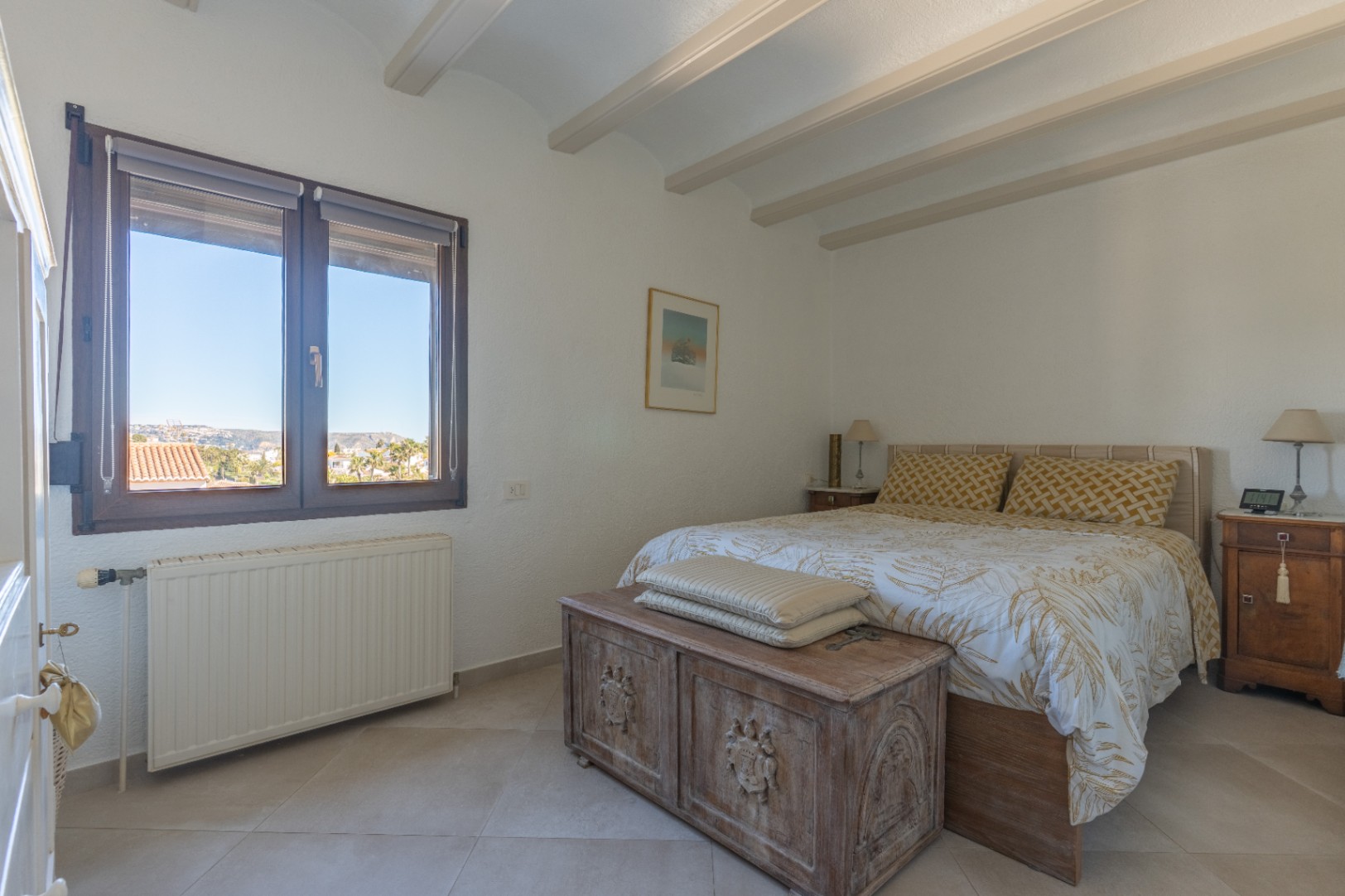 Dormitorio con vigas de madera a la vista, suelos cerámicos y un baúl rústico tallado. Incluye calefacción central y ventana con vistas al entorno de Jávea.