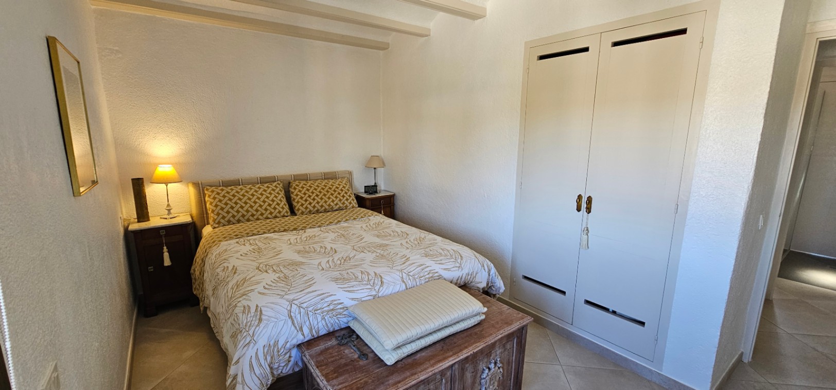 Dormitorio con vigas vistas en el techo, armario empotrado de puertas blancas y un baúl de madera rústico a los pies de la cama.