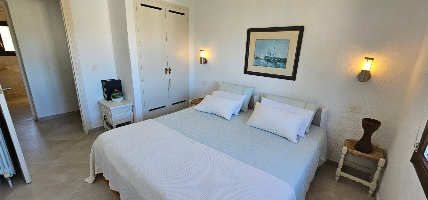 Dormitorio luminoso con cama doble, armarios empotrados blancos y suelos de gres. Incluye apliques de pared de luz cálida y acceso directo al pasillo.