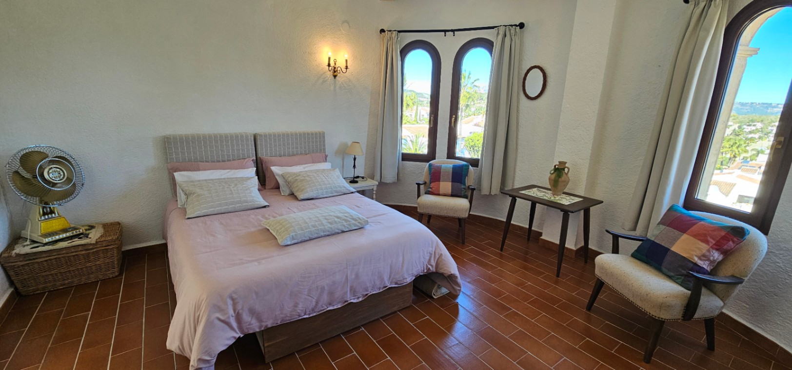Dormitorio luminoso con suelo de terracota, ventanas de arco tradicionales y mobiliario clásico. Incluye zona de estar con sillones y vistas despejadas al entorno de Jávea.