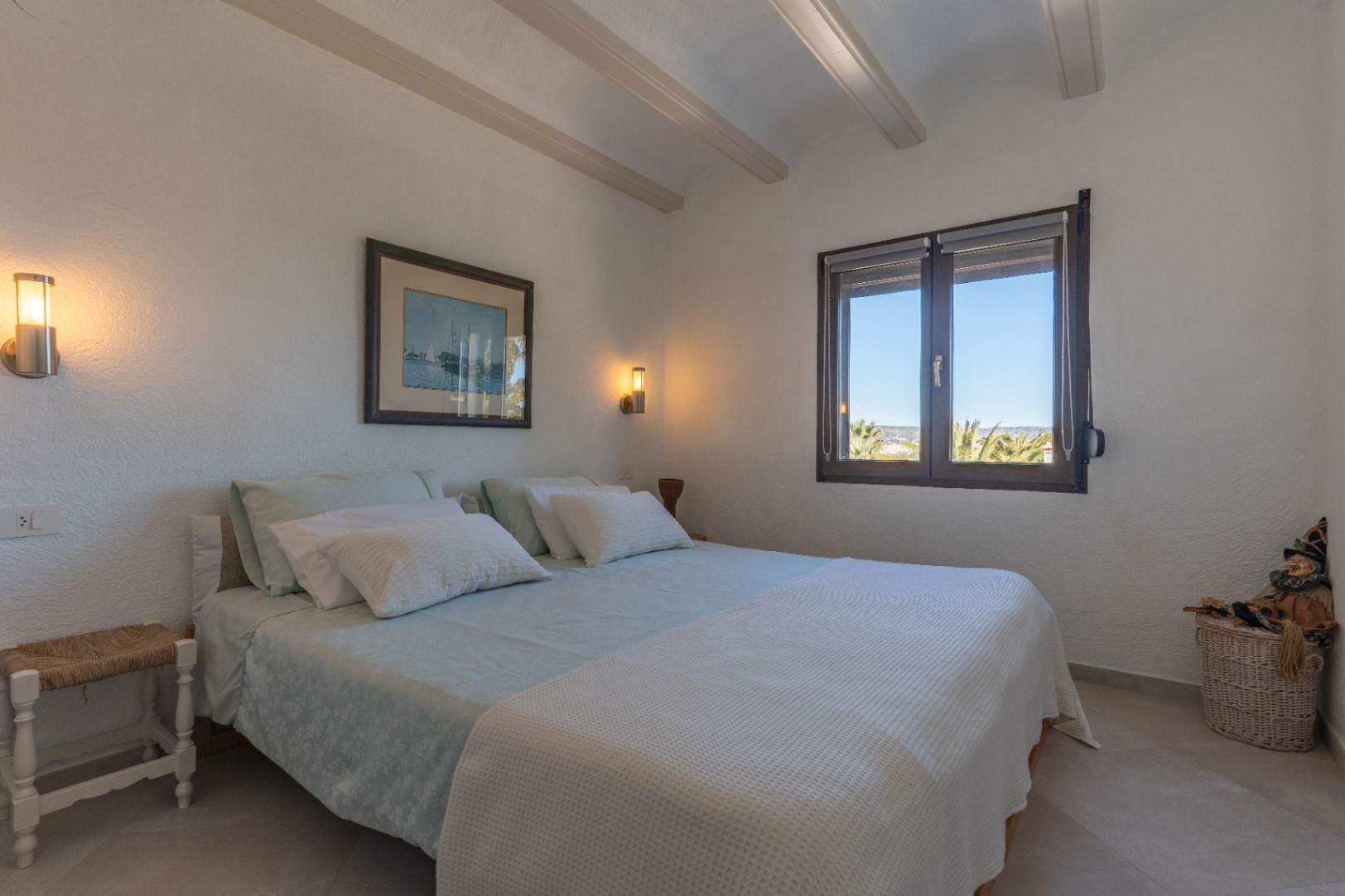 Dormitorio luminoso con vigas de madera a la vista, apliques de pared modernos y una ventana con marco de madera que ofrece vistas al jardín mediterráneo.
