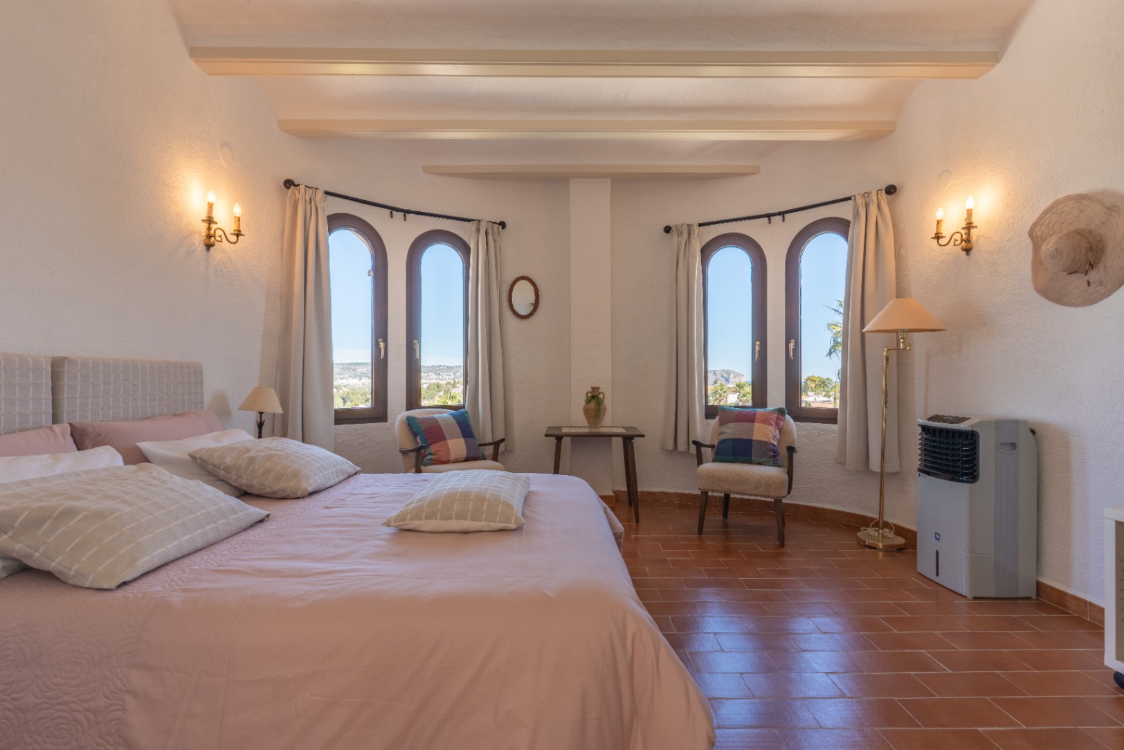 Dormitorio luminoso con vigas vistas, suelo de terracota y ventanas de arco que ofrecen vistas despejadas hacia Jávea, complementado con apliques de pared clásicos.