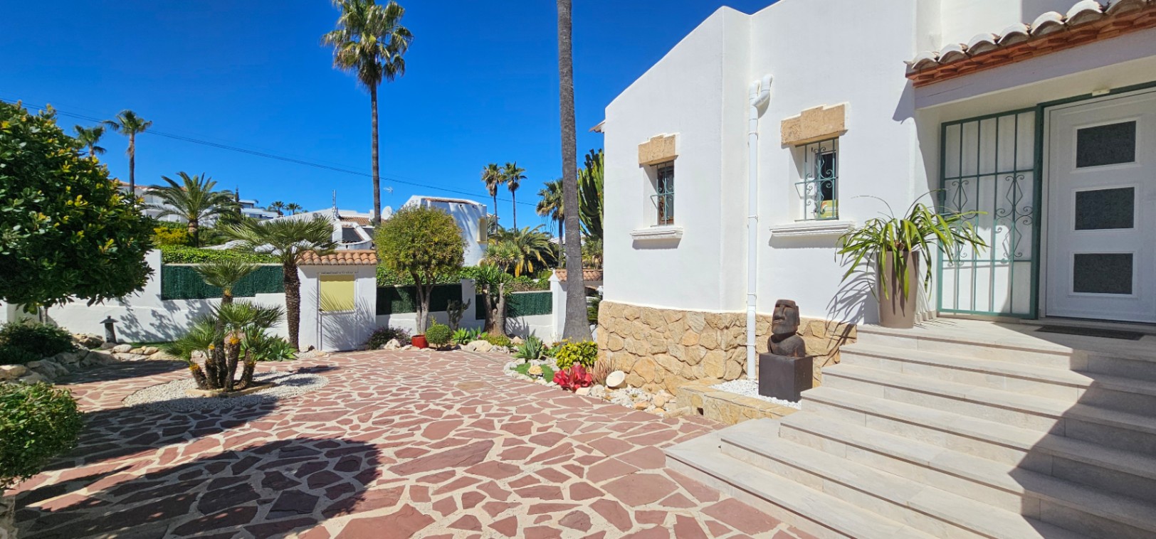 Entrada principal con escalinata blanca y fachada mediterránea. Destaca el pavimento de piedra irregular y el jardín ornamental con palmeras bajo el cielo despejado de Jávea.