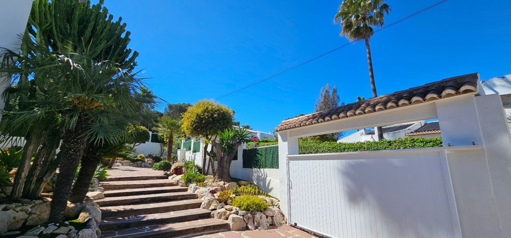 Entrada principal con puerta corredera blanca y escalinata de piedra natural rodeada de vegetación mediterránea, incluyendo cactus y palmeras bajo un cielo despejado.