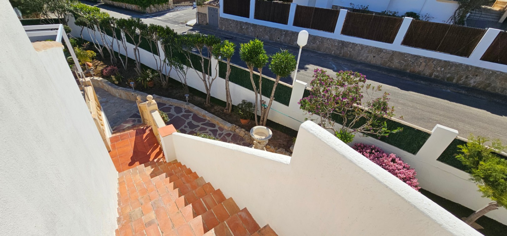 Escalera exterior de baldosas cerámicas que desciende hacia un jardín cuidado con senderos de piedra natural y vegetación mediterránea perimetral.