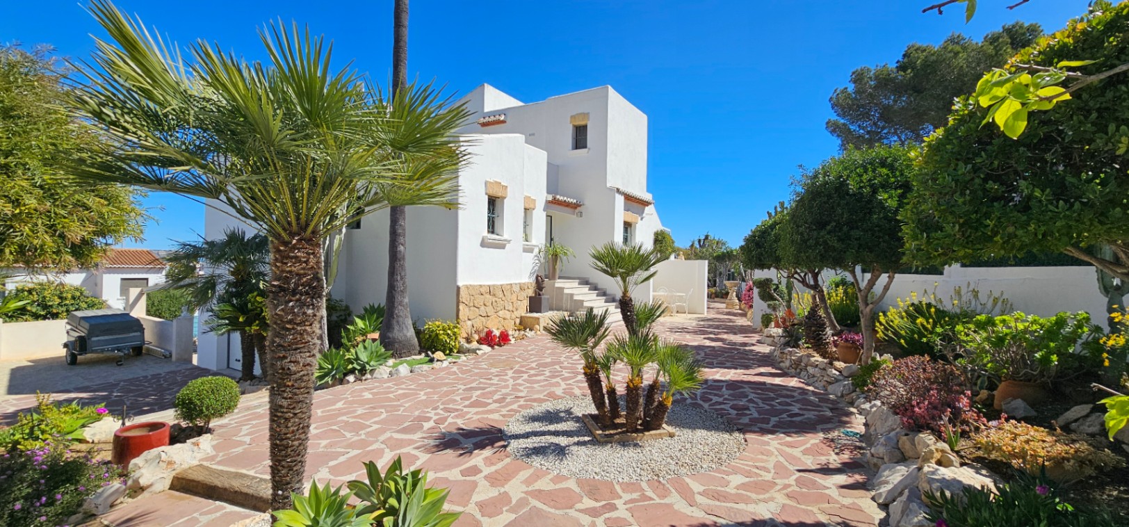 Exterior de la villa con fachada blanca y zócalo de piedra, rodeada de un jardín mediterráneo con palmeras y pavimentación de piedra irregular.
