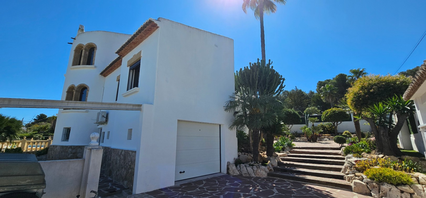 Fachada exterior blanca con base de piedra natural, garaje integrado y escalinata de piedra rodeada de vegetación mediterránea y palmeras.