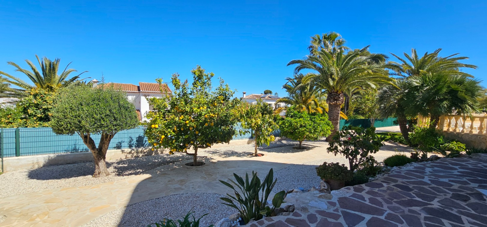 Jardín mediterráneo con árboles frutales, olivos y palmeras, rodeado de pavimentos de piedra irregular y zonas de grava bajo un cielo despejado.